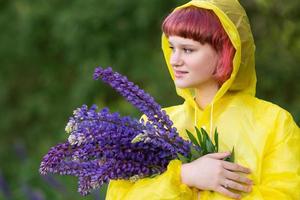 A cute teenage girl with pink hair and a bouquet of lupines is standing in a yellow raincoat photo