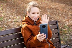 The boy is talking by video on the phone, sitting on a bench and laughing photo