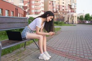 The girl is sitting on a bench, laughing and looking at the phone, there is a laptop nearby photo