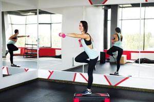 The trainer stands on the step, stretching his arm forward with dumbbells, pulling his leg back. photo