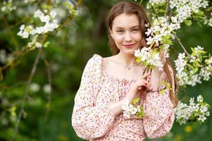 A girl in a pink dress stands near a white tree with flowers looks at the camera and smiles photo
