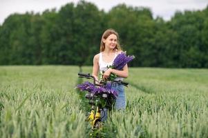 A cute girl is standing with a bouquet of lupines in a field near a bicycle photo