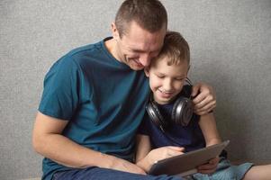 Dad hugs his son sitting on the floor with big headphones, playing in the gadget photo