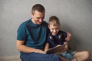 Dad hugs his son sitting on the floor with big headphones, playing in the gadget photo