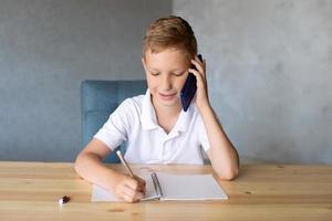 Cute boy writes in an open notebook and looks at the phone. photo