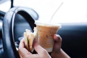 Asian woman driver hold ice coffee cup and sandwich bread for eat and drink in car, dangerous and risk an accident. photo