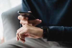 hombre utilizando teléfono inteligente y reloj inteligente para rastreo actividad en sofá en vivo habitación a hogar, inalámbrico conexión Entre el reloj y móvil teléfono. foto