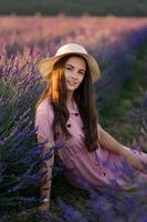 A cheerful young girl in a pink dress and a straw hat in her hands stands among lavender bushes. Sunset. photo