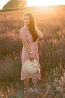 A cheerful young girl in a pink dress and a straw hat in her hands stands among lavender bushes. Sunset. photo