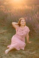 un alegre joven niña en un rosado vestir y un Paja sombrero en su manos soportes entre lavanda arbustos puesta de sol. foto