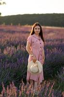 A cheerful young girl in a pink dress and a straw hat in her hands stands among lavender bushes. Sunset. photo