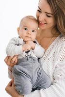 un joven madre sostiene su pequeño hijo en su brazos y sonrisas en un blanco antecedentes foto