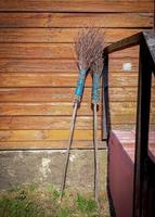 Two old brooms with blue rope holding all branches together standing up against the wall of wooden plank house with iron railing on sunny day photo