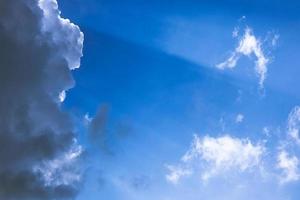 White soft clouds slowly being covered by one big dark cloud from left side while rays of light are coming out from behind of it on blue sky photo