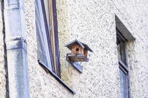 Handmade wooden bird feeder little house with round hole on yellow brick wall of a building with open window photo