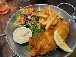 cerca arriba ver de delicioso pescado y papas fritas con francés papas fritas, César ensalada con balsámico salsa y sarro salsa o mayonesa foto