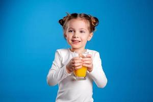 little girl with a funny hairstyle in a white T-shirt drinks juice from a glass. blue background. useful products for children. space for text. High quality photo