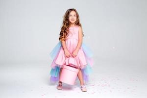 hermosa pequeño niña en un elegante rosado vestir poses sentado en un gris antecedentes y lanza papel picado. Moda y estilo. espacio para texto. foto en el estudio. alto calidad foto