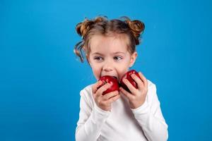 cute little girl with curls in a striped dress is holding a big red apple in her hands. blue background. useful products for children. healthy snack. space for text. High quality photo