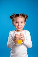 pequeño niña con un gracioso peinado en un blanco camiseta bebidas jugo desde un vaso. azul antecedentes. útil productos para niños. espacio para texto. alto calidad foto