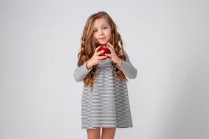 cute little girl with curls in a striped dress is holding a big red apple in her hands. blue background. useful products for children. healthy snack. space for text. High quality photo