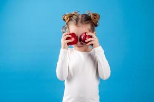 linda pequeño niña con chinos en un a rayas vestir es participación un grande rojo manzana en su manos. azul antecedentes. útil productos para niños. sano bocadillo. espacio para texto. alto calidad foto