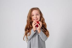 linda pequeño niña con chinos en un a rayas vestir es participación un grande rojo manzana en su manos. azul antecedentes. útil productos para niños. sano bocadillo. espacio para texto. alto calidad foto