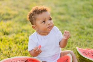 a little swarthy girl in white clothes eats a watermelon on a green lawn. picnic in the park. happy childhood. space for text. High quality photo