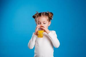 pequeño niña con un gracioso peinado en un blanco camiseta bebidas jugo desde un vaso. azul antecedentes. útil productos para niños. espacio para texto. alto calidad foto