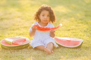 un pequeño moreno niña con Rizado pelo en un pálido rosado vestir come un sandía en el césped. picnic en el parque. contento infancia. espacio para texto. alto calidad foto