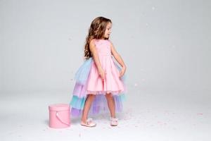 hermosa pequeño niña en un elegante rosado vestir poses sentado en un gris antecedentes y lanza papel picado. Moda y estilo. espacio para texto. foto en el estudio. alto calidad foto