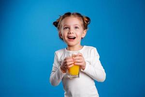 pequeño niña con un gracioso peinado en un blanco camiseta bebidas jugo desde un vaso. azul antecedentes. útil productos para niños. espacio para texto. alto calidad foto