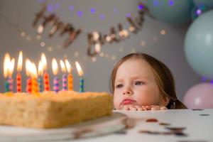 cute little girl blows out candles on a birthday cake at home against a backdrop of balloons. Child's birthday photo