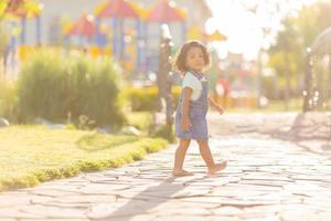 retrato pequeño contento juguetón niñito moreno niña en un mezclilla vestido de verano en pie en el jardín en un soleado día. caminando en el Fresco aire. concepto de un contento infancia. espacio para texto. alto calidad foto