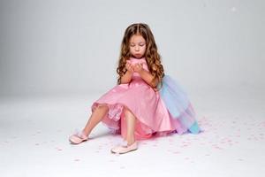 hermosa pequeño niña en un elegante rosado vestir poses sentado en un gris antecedentes y lanza papel picado. Moda y estilo. espacio para texto. foto en el estudio. alto calidad foto