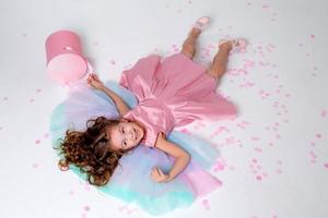 beautiful little girl in a chic pink dress lies on the floor strewn with confetti. top view. fashion and style. child celebrates his birthday. photo in the studio. space for text. High quality photo