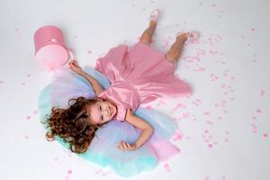 hermosa pequeño niña en un elegante rosado vestir poses sentado en un gris antecedentes y lanza papel picado. Moda y estilo. espacio para texto. foto en el estudio. alto calidad foto