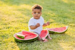 a little swarthy girl in white clothes eats a watermelon on a green lawn. picnic in the park. happy childhood. space for text. High quality photo