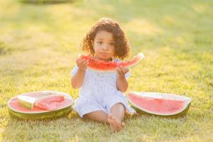 a little swarthy girl with curly hair in a pale pink dress eats a watermelon on the lawn. picnic in the park. happy childhood. space for text. High quality photo