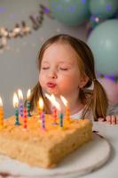 cute little girl blows out candles on a birthday cake at home against a backdrop of balloons. Child's birthday photo