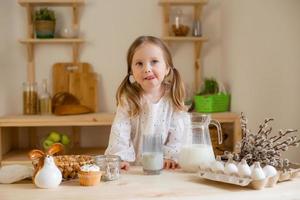 un linda pequeño niña bebidas Leche a hogar en un de madera cocina. Leche día foto