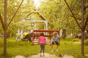 Two cute little sisters having fun on a swing together in beautiful summer garden on warm and sunny day outdoors. Active summer leisure for kids. photo