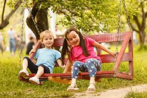 Two cute little sisters having fun on a swing together in beautiful summer garden on warm and sunny day outdoors. Active summer leisure for kids. photo