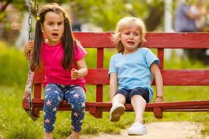 Two cute little sisters having fun on a swing together in beautiful summer garden on warm and sunny day outdoors crying photo