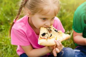 pequeño niña con su ojos amplio abierto con deleite comiendo Pizza en el verano parque foto