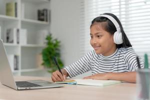 online tutor online course for Asian children. video conference on laptop at home. play back online course, girl student wear earphones to study, notes homework, video classes, new education. photo