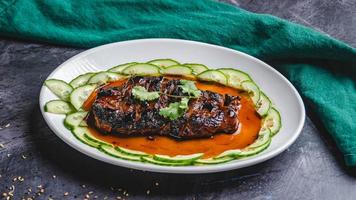 Grilled chicken breast covered with sauce, cucumbers, and coriander on a white plate. photo