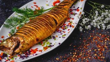 Mouth-watering whole stuffed fish served on a white plate with ginger, tomato, coriander, gypsophila paniculata on the side, and crushed red pepper flakes. photo