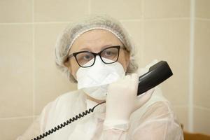 un mayor hembra médico en un médico máscara respuestas teléfono llamadas durante un epidemia o pandemia. foto