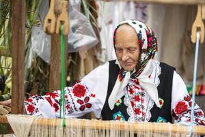 An old Belarusian or Ukrainian woman in an embroidered shirt at a vintage loom. Slavic elderly woman in national ethnic clothes. photo